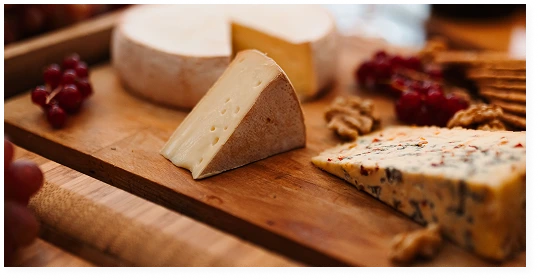 A wooden platter with a selection of cheeses, nuts, redcurrants and a glass of white wine