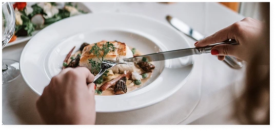A man is eating a baked fish fillet with mushrooms with a knife and fork at a set restaurant table