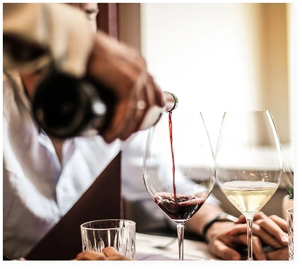 A waiter is pouring red wine from a bottle into a glass on a laid table