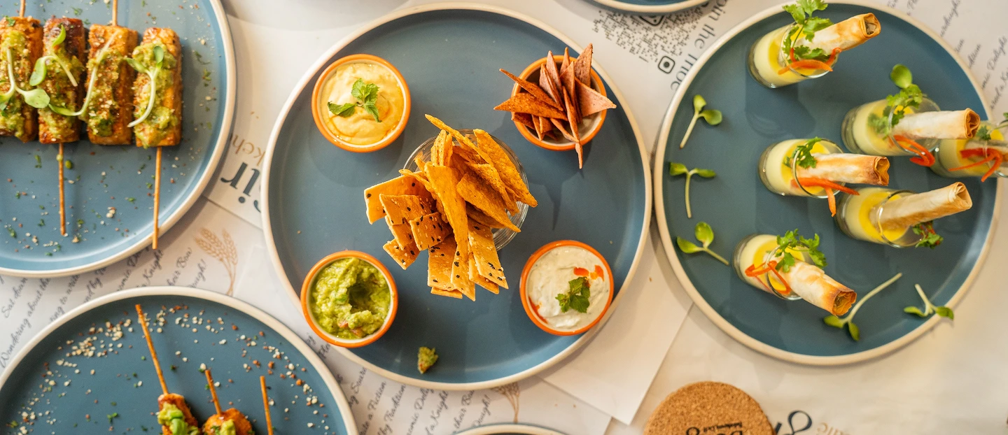 A top-down view of a table with a variety of snacks, nacho crisps and dips on blue plates